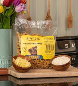 Bag of Betty Miller Cheese Little Bones dog food on a wooden surface with bowls of ingredients and flowers.