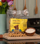 Bag of Betty Miller Everyday Liver Bones dog treats on a wooden surface with a bowl of treats and flowers in the background.