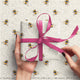 Gift wrapped in paper with bee pattern and pink ribbon, held by hands against matching background.